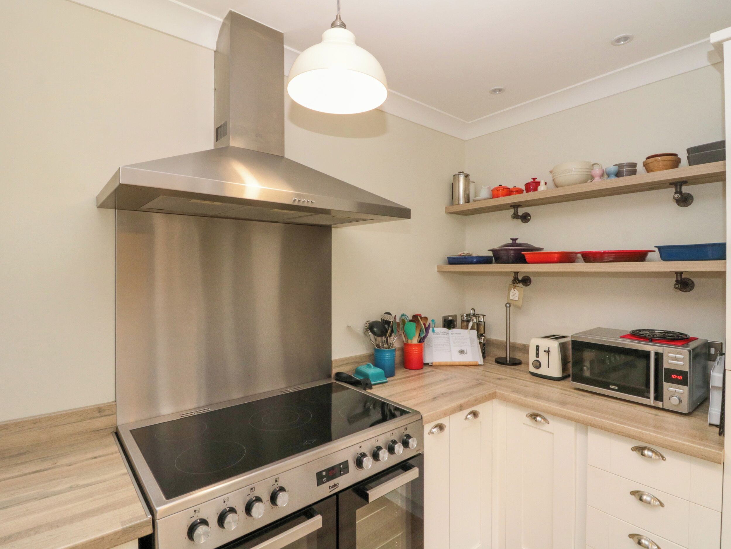 Una cucina moderna con una cappa in acciaio inossidabile, forno integrato, microonde e tostapane. Sono presenti mobili in legno e un piano di lavoro con diversi utensili e elettrodomestici da cucina.