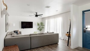 A modern living room with a grey sectional sofa, a flat-screen TV, a ceiling fan, and a large window with white curtains. - Chic Scottsdale Stay | Pool, Hot Tub! (Scottsdale)