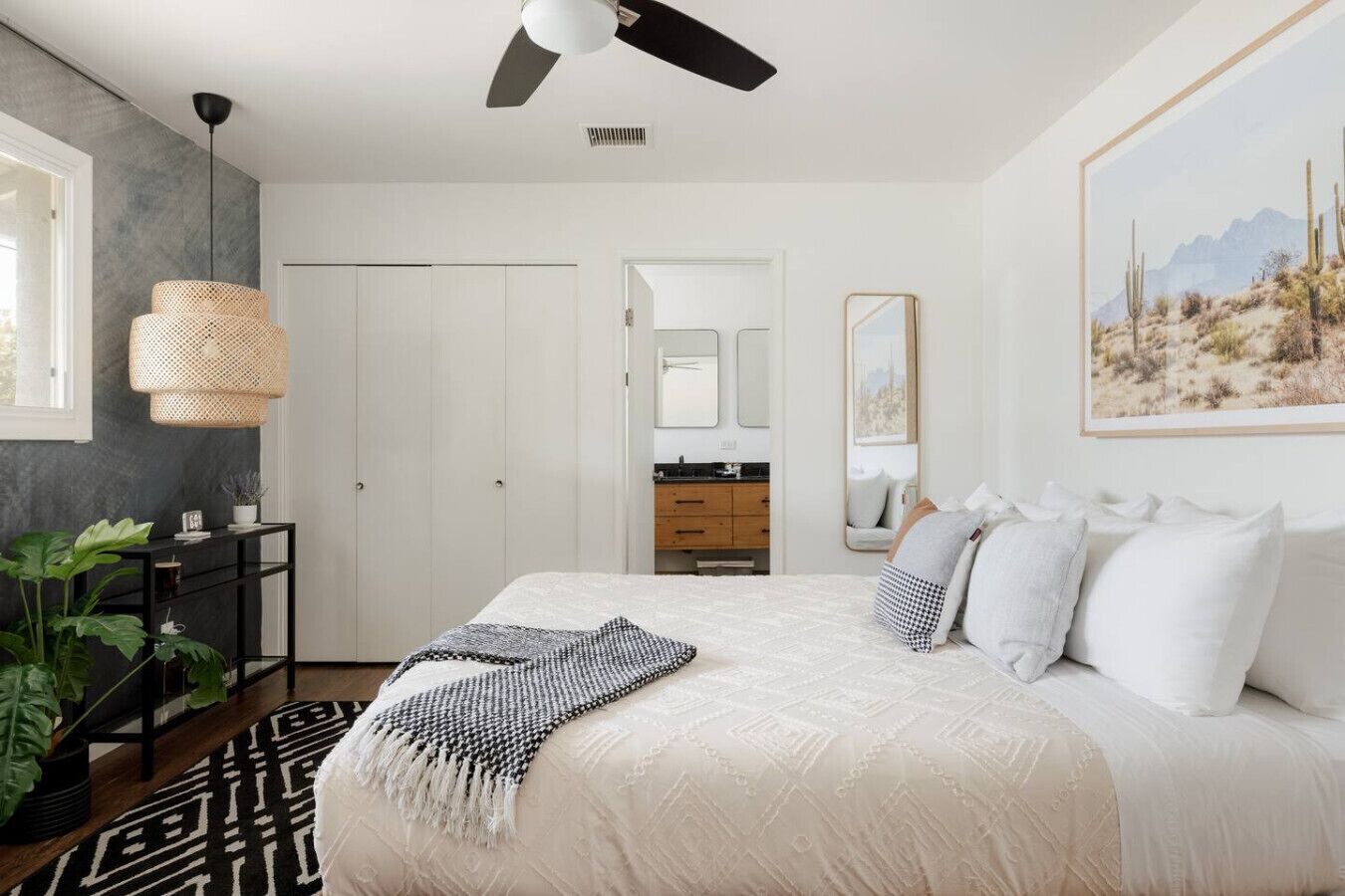 A bedroom with a large bed, a ceiling fan, a black console table with a plant, and a painting of a desert landscape.