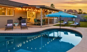 Un área junto a la piscina con sillas de descanso, una parrilla y luces colgantes.