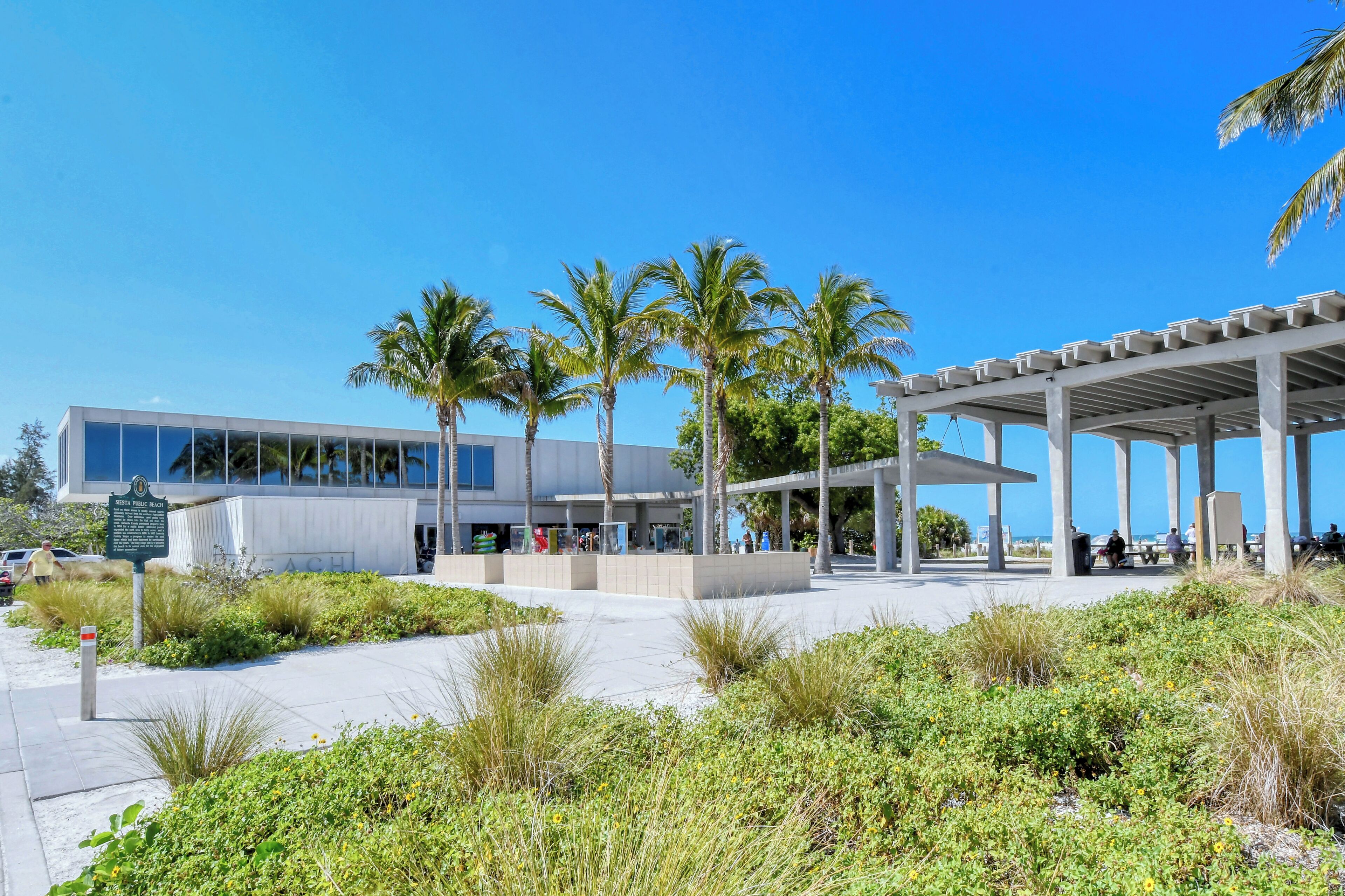 A modern building with a glass facade, surrounded by palm trees and a covered outdoor seating area.