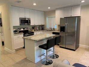 A modern kitchen with white cabinets, stainless steel appliances, a granite countertop, and two bar stools. - Casa Eli - Lovely Cottage (Sarasota)