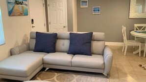 Un salon avec un canapé d’angle beige, deux coussins bleu foncé, une porte blanche, une petite table à manger avec des chaises blanches et un tableau représentant une scène de plage accroché au mur.