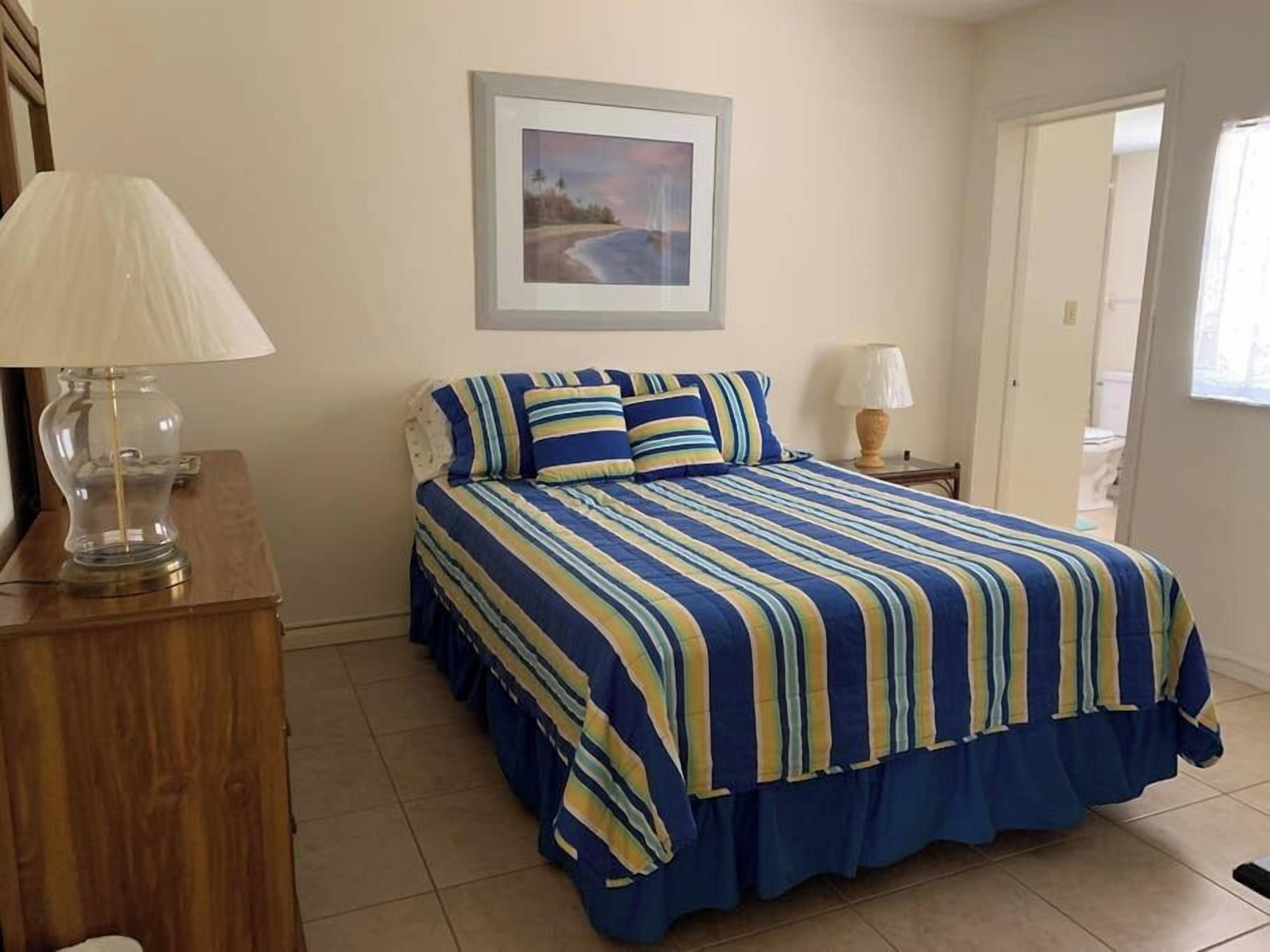 Un dormitorio con una cama, un aparador de madera, un cuadro enmarcado en la pared y una puerta que da a otra habitación.