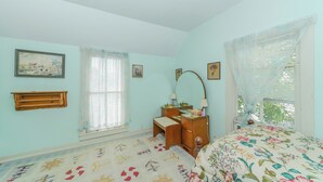 Un dormitorio con cama, cómoda, espejo, ventana con cortinas y estante fijado a la pared.