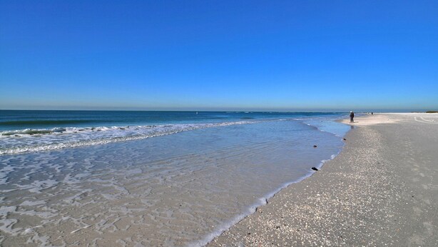 Une vaste plage de sable fin, avec de douces vagues, un ciel bleu clair et quelques personnes au loin.