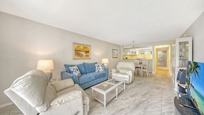 A modern living room with a blue sofa, white armchairs, a coffee table, and a dining area with a white table and chairs.