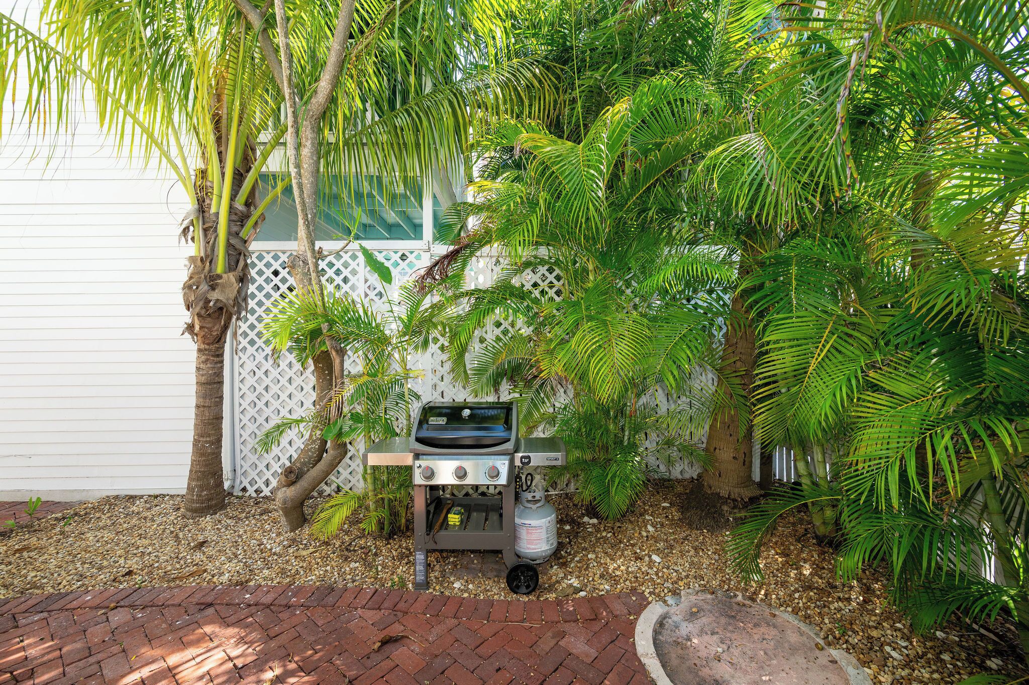 Een draagbare grill is opgezet in een tuin met palmbomen en een witte omheining op de achtergrond.