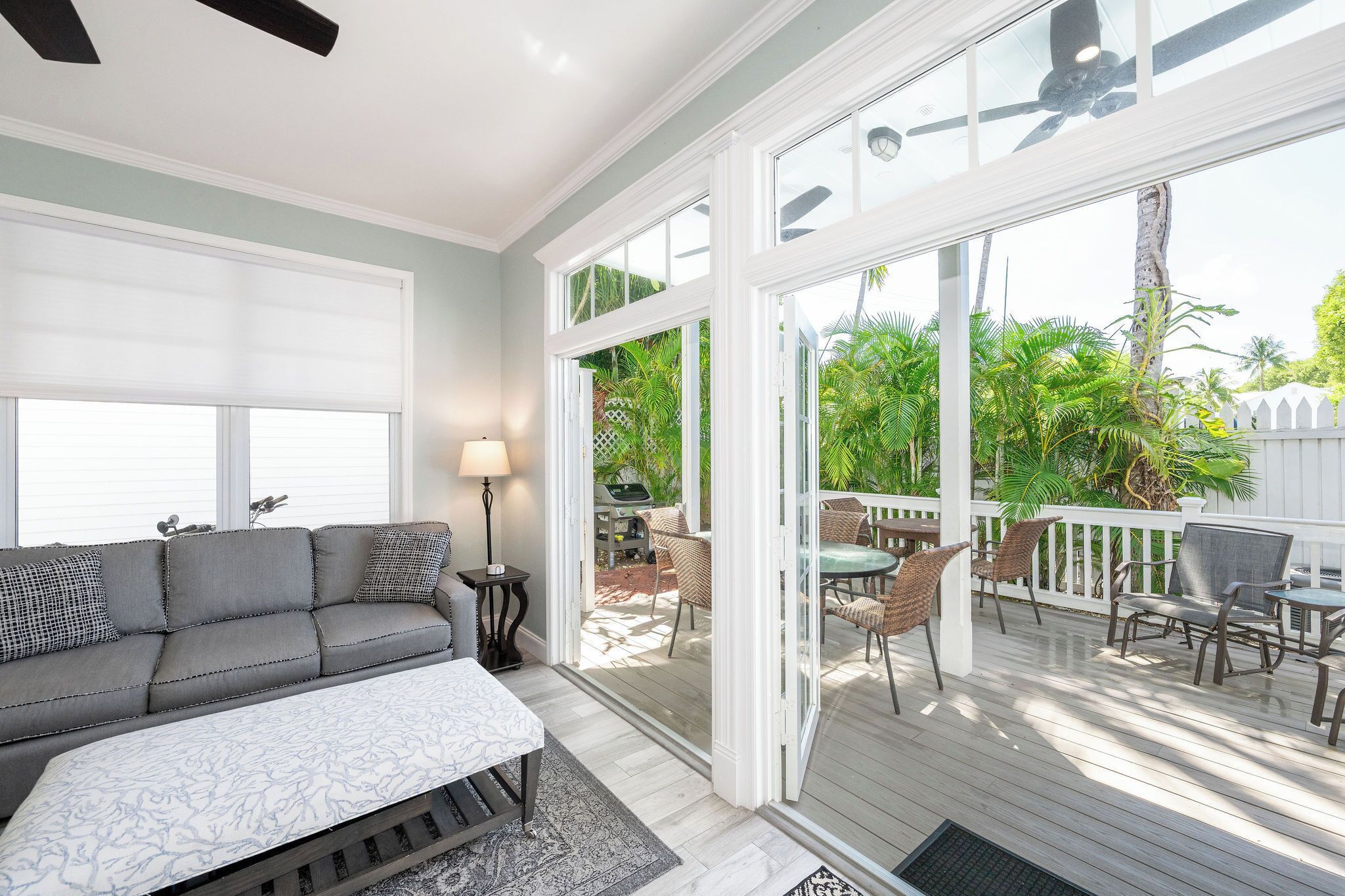 A spacious living room with a gray sofa, a white coffee table, and a glass door leading to an outdoor patio with wicker furniture and a view of greenery.
