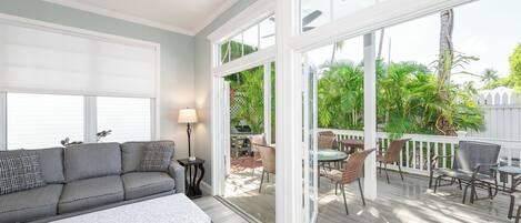 A spacious living room with a gray sofa, a white coffee table, and a glass door leading to an outdoor patio with wicker furniture and a view of greenery.