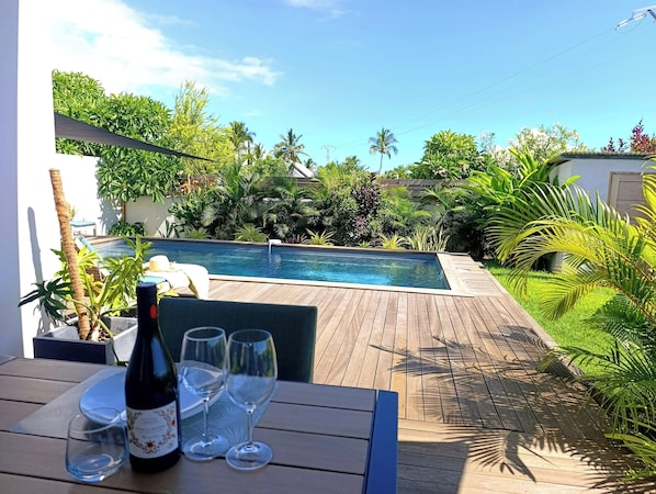 Une terrasse en bois avec une piscine, entourée d’une végétation luxuriante et sous un ciel d’un bleu éclatant.