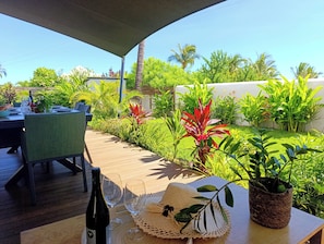 Un espace extérieur aménagé pour les repas, avec une terrasse en bois, une table dressée avec une bouteille de vin, des verres et un chapeau.