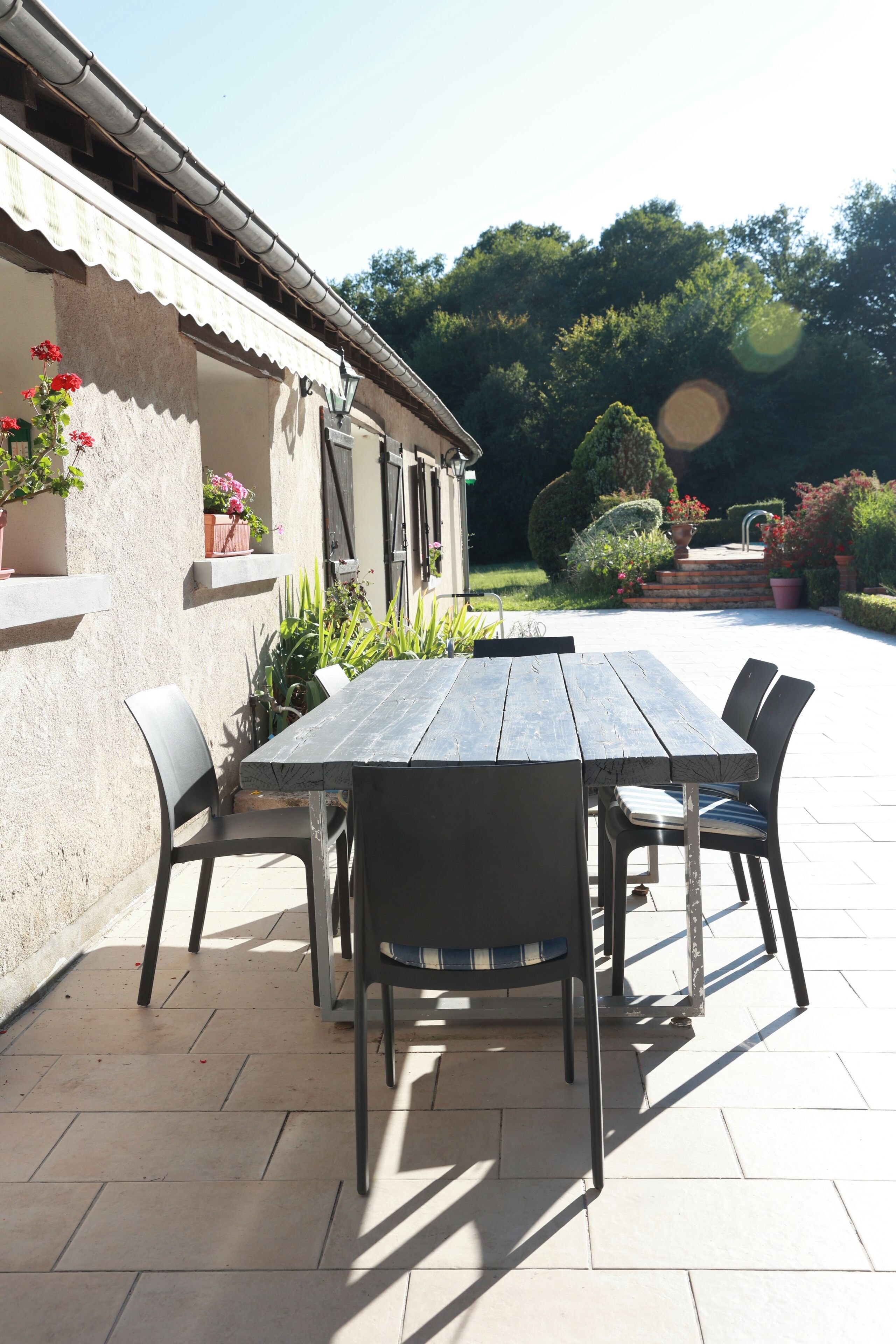 Een terras buiten met een houten tafel en stoelen, omgeven door potplanten en een tuin.