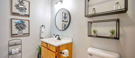 A bathroom with a wooden vanity, a white sink, a mirror, and framed artwork on the wall.