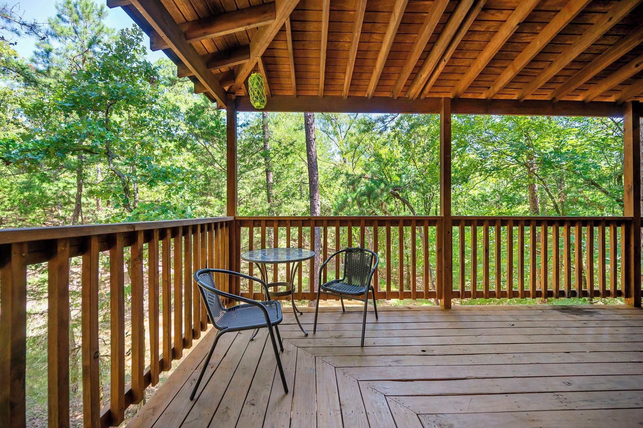 A wooden deck with two metal chairs and a small table, surrounded by a wooden railing, overlooking a forested area.
