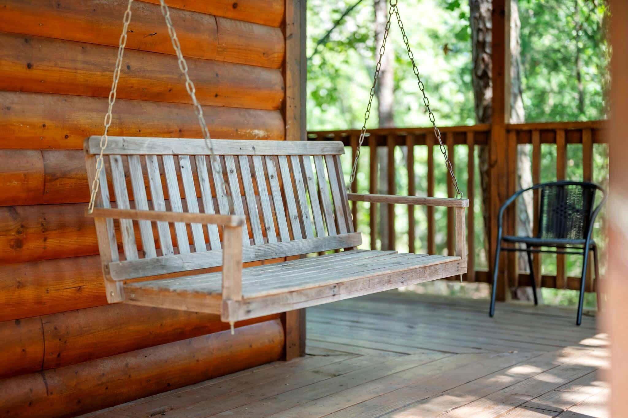 A wooden bench hanging from a chain, a metal chair, and a wooden deck with a wooden fence in the background.