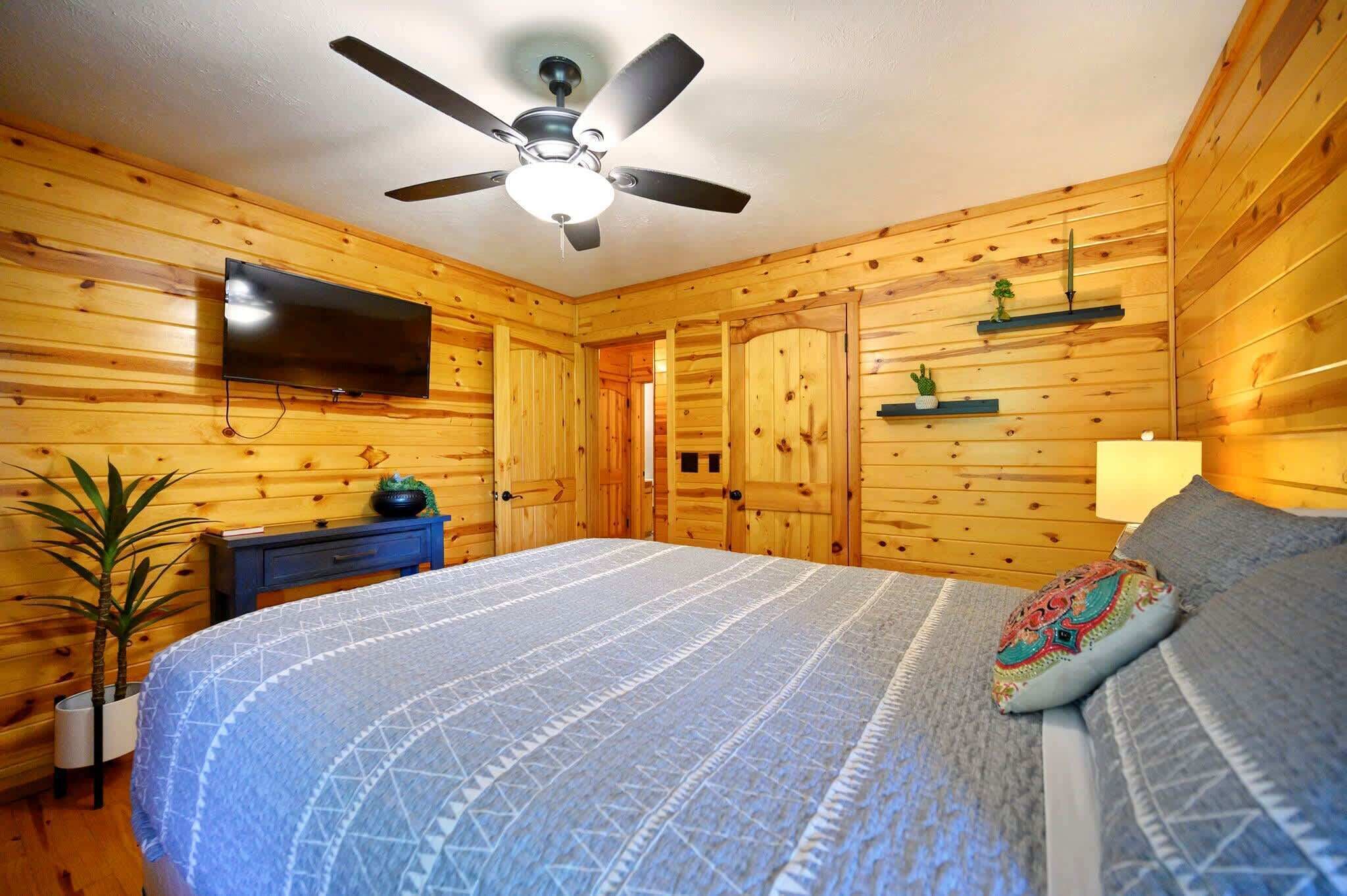 A wooden-paneled room with a bed, a ceiling fan, a flat-screen TV, a nightstand, a potted plant, and a shelf with decorative items.