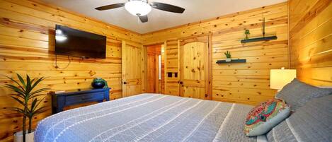 A wooden-paneled room with a bed, a ceiling fan, a flat-screen TV, a nightstand, a potted plant, and a shelf with decorative items.