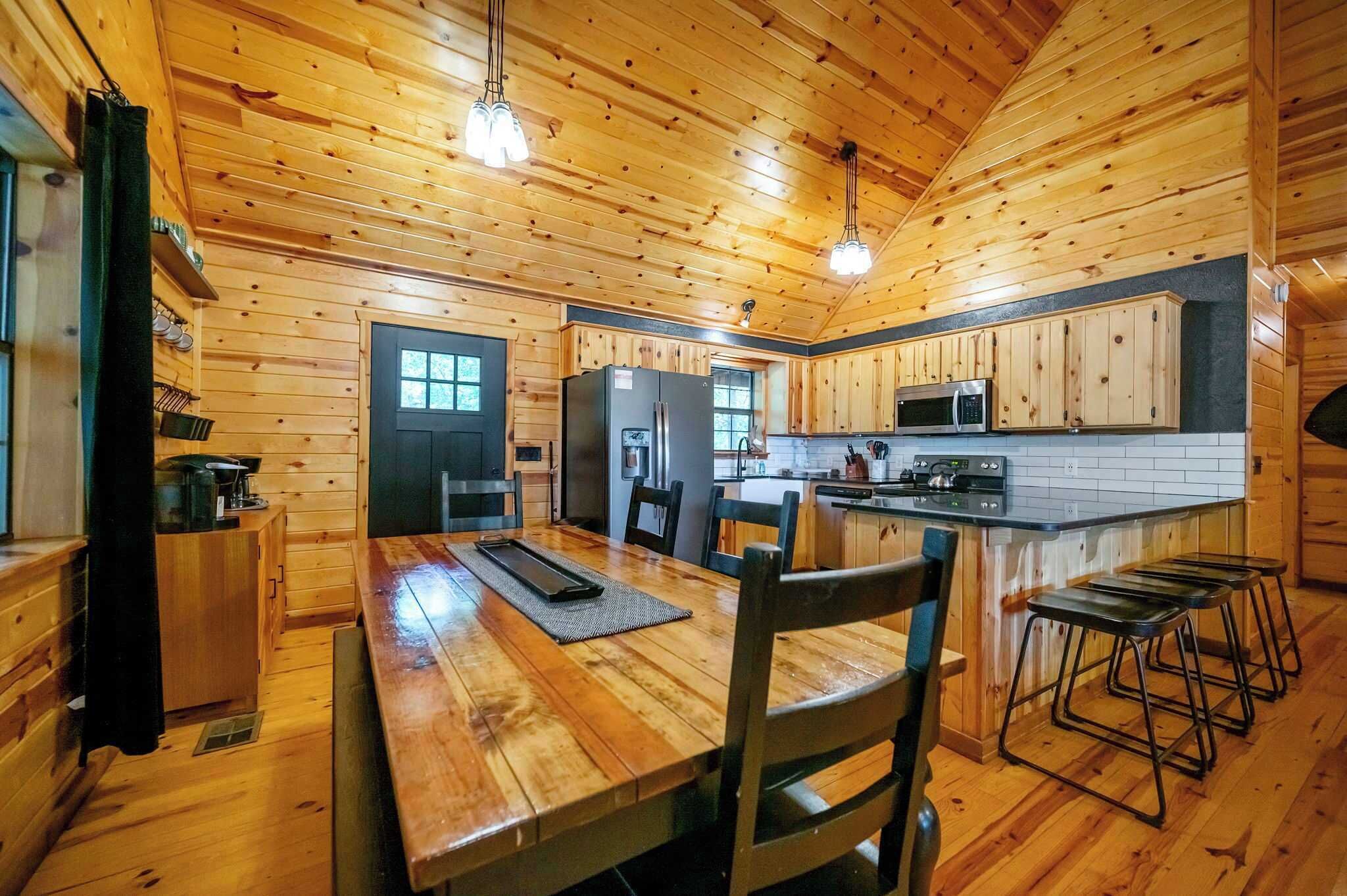 A wooden cabin interior with a dining area, kitchen, and bar stools.