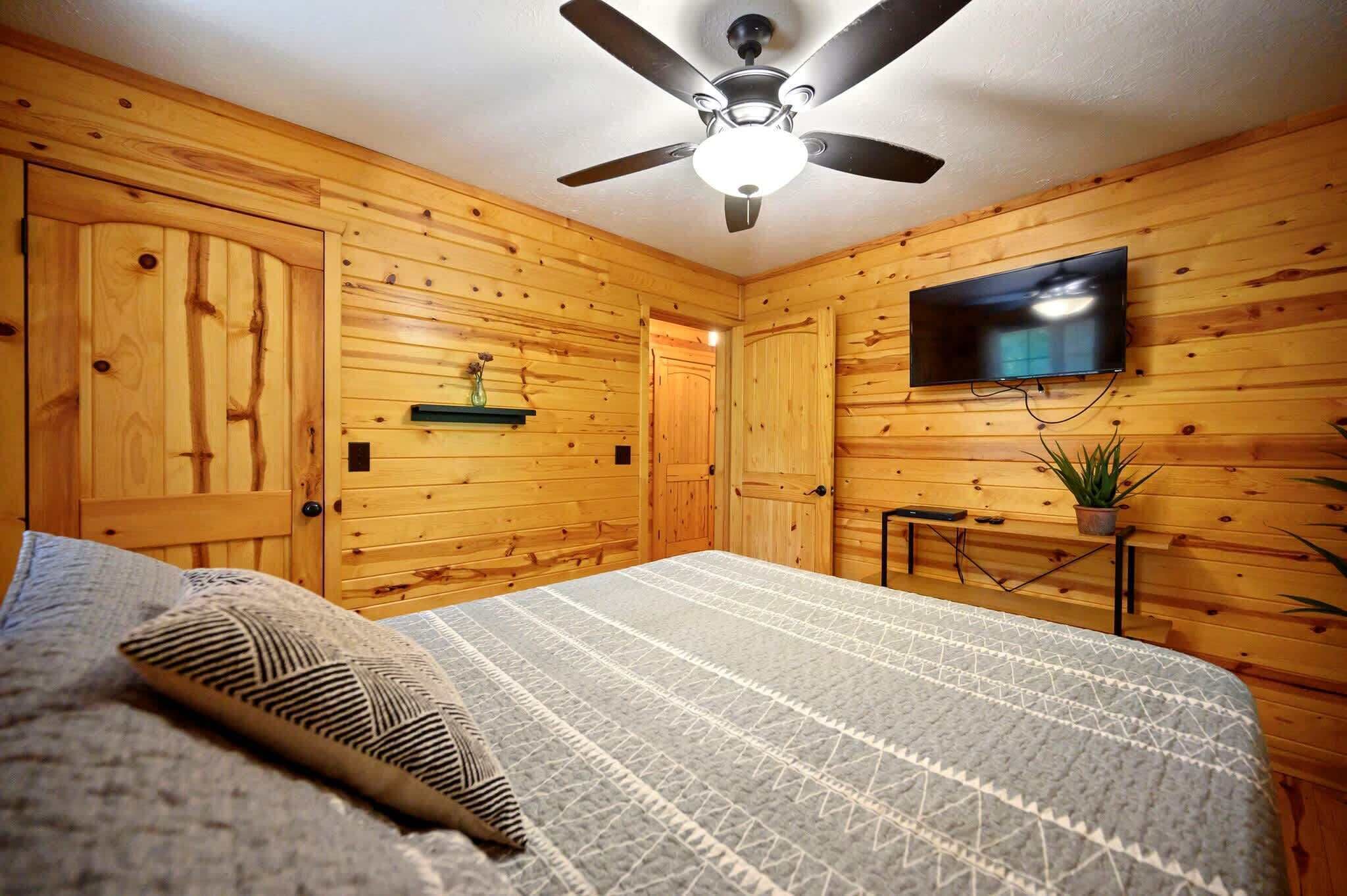 A bedroom with wooden walls, a ceiling fan, a flat-screen TV mounted on the wall, and a bed with a patterned blanket.