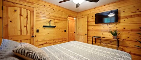 Un dormitorio con paredes de madera, ventilador de techo, televisor de pantalla plana montado en la pared y una cama con manta estampada.