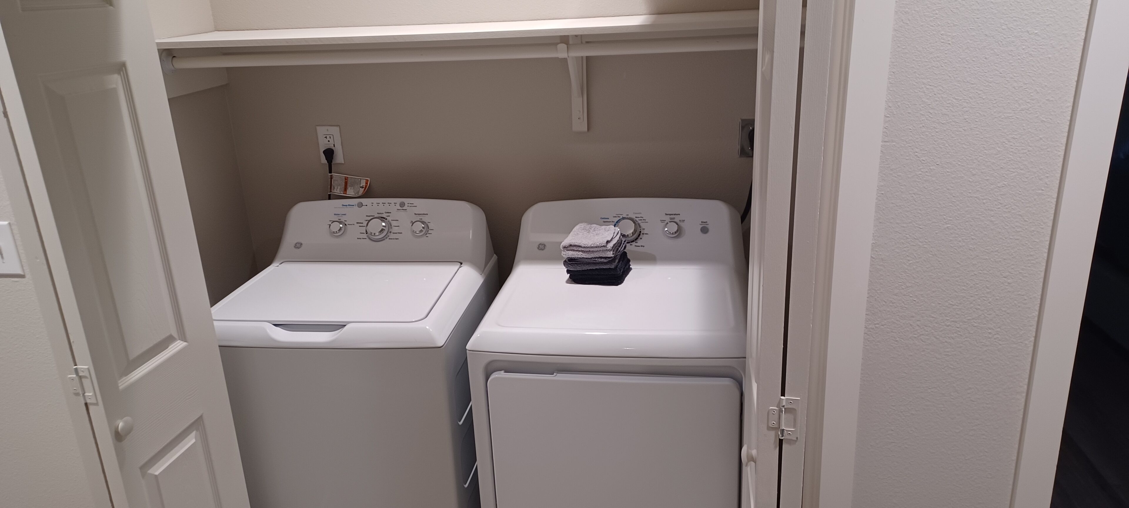 Un cuarto de lavado con dos lavadoras blancas y una secadora, un enchufe y un estante sobre las máquinas.