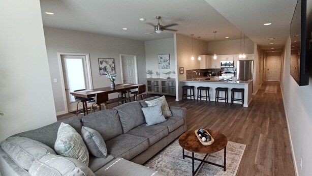 A modern living room with a grey sofa, a coffee table, and a dining area with a wooden table and chairs. A kitchen with a central island and bar stools is visible in the background. - Charming 2-Bedroom Apartment in Downtown Manor (Manor)