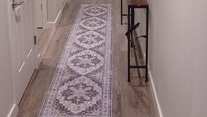 A hallway with wooden flooring, white walls, and a patterned rug. There is a door at the end and a small table with a picture frame on the right. - Charming 2-Bedroom Apartment in Downtown Manor (Manor)