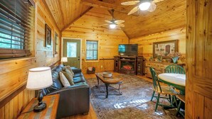 A cozy log cabin interior with a fireplace, a sofa, a dining table, and a ceiling fan. - Sunset Ridge Cozy Cabin for 6 (Broken Bow)
