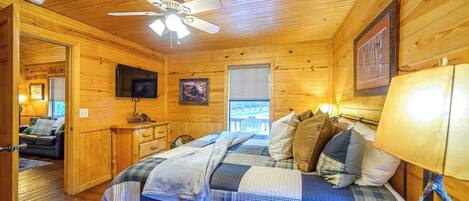 A cozy cabin room with a bed, a ceiling fan, a TV, and a window.