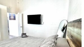 Un dormitorio con cama, ventilador de techo, televisor montado y puerta que da a otra habitación.