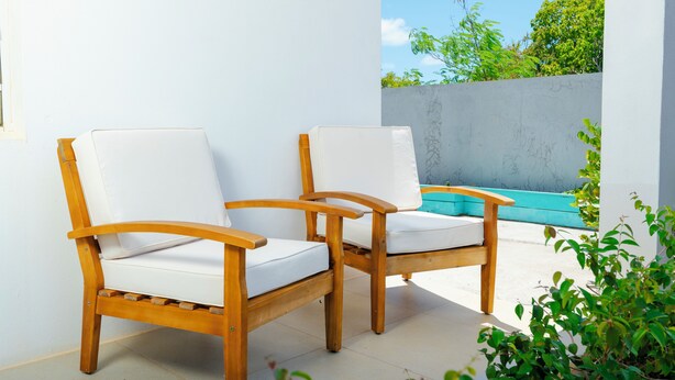 Deux chaises en bois avec des coussins blancs, placées dans un espace extérieur ensoleillé, doté d’un sol carrelé et où l’on aperçoit une piscine bleue à l’arrière-plan.