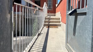 Une ruelle étroite, avec un immeuble rouge, une clôture métallique blanche et un escalier en béton menant à une porte.