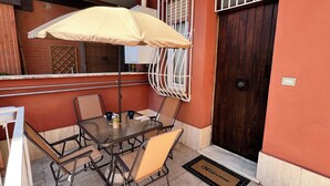 Une terrasse extérieure avec une table et des chaises, un parasol beige et une porte avec un paillasson.