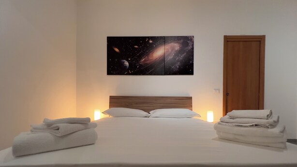 Un lit bien fait, avec du linge de lit blanc et des oreillers, une tête de lit en bois et, au-dessus, une œuvre d’art murale sur le thème de la galaxie.