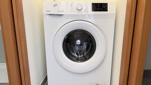 Vue de face d’un lave-linge à chargement frontal blanc, doté d’un panneau de commande sur le dessus, situé dans une pièce dont le sol est recouvert de carreaux.