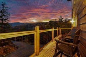 Eine Holzterrasse mit Geländer, mit Blick auf eine bewaldete Landschaft und einem Sonnenuntergang im Hintergrund.