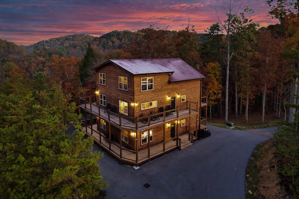 A two-story wooden cabin with a balcony, surrounded by a forested area.