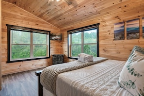 Un dormitorio de cabaña de madera con una cama, dos ventanas, un aparador y un televisor.