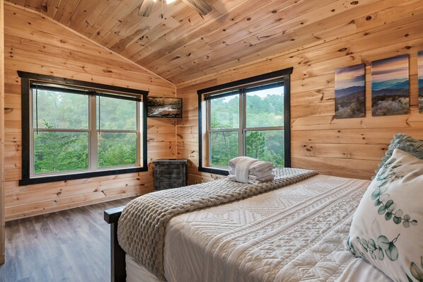 Un dormitorio de cabaña de madera con una cama, dos ventanas, un aparador y un televisor.