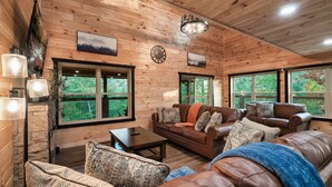 A cozy cabin interior with wooden walls, a leather sofa, a coffee table, and large windows offering a view of the forest.