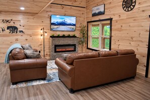A cozy cabin interior with a fireplace, leather sofas, a television, and wooden walls. - Spacious Mountain Cabin with Hot Tub, Sleeps 12 (Pigeon Forge)