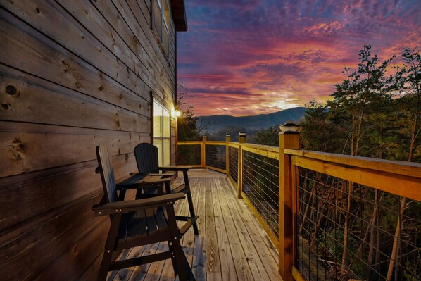 Una terraza de madera con una silla y una mesa, con vistas a un paisaje boscoso al atardecer.