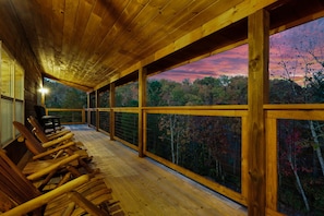 Una terraza de madera con sillas orientadas a una vista panorámica de árboles y un atardecer.