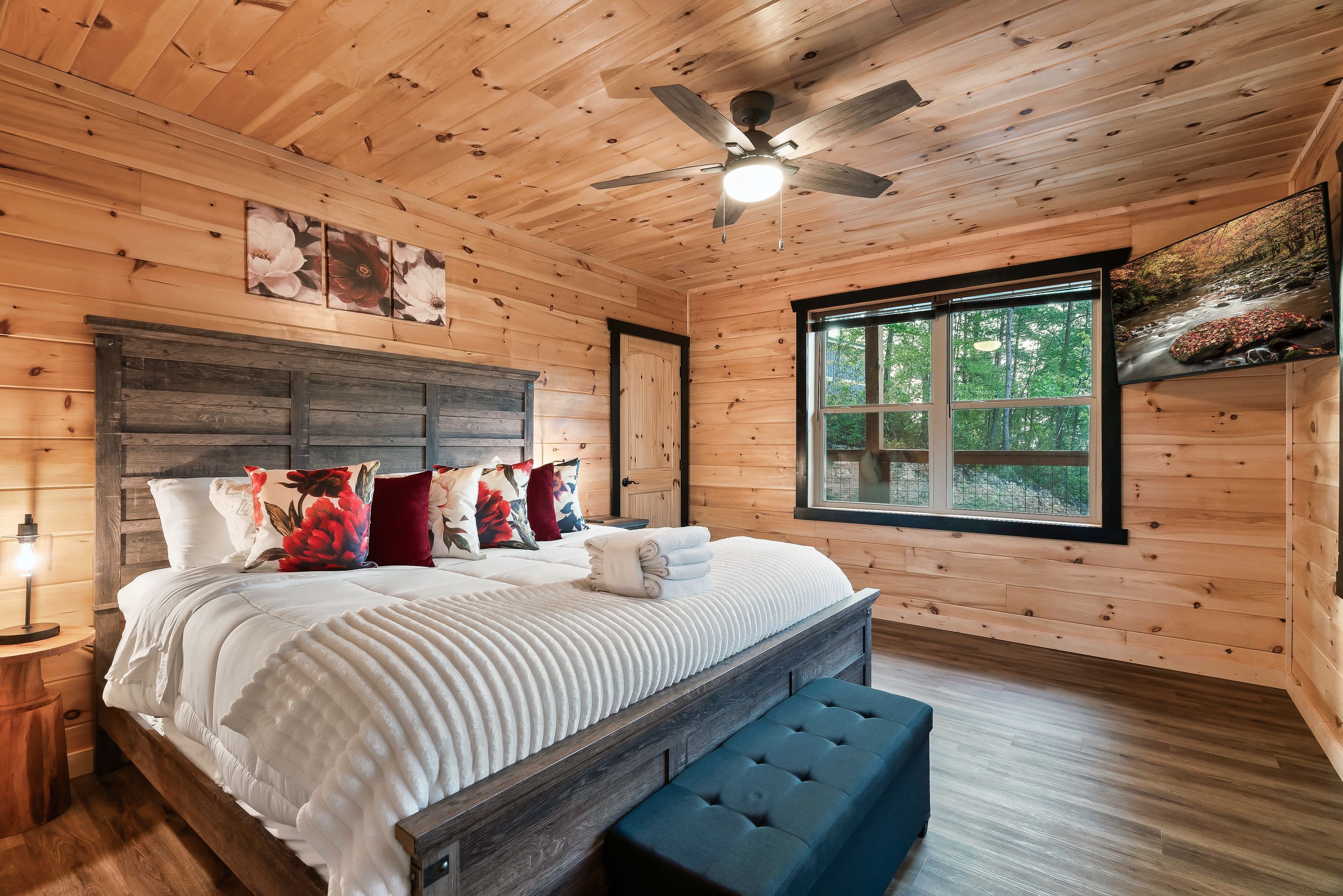 A wooden cabin bedroom with a bed, pillows, a bench, a ceiling fan, and a window with a view of a forest.