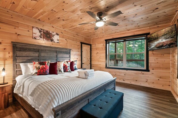 Dormitorio de cabaña de madera con una cama, almohadas, un banco, un ventilador de techo y una ventana con vistas a un bosque.