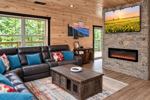 A modern living room with a leather sofa, a coffee table, a fireplace, and a large window with a view of trees. - Spacious Mountain Cabin with Hot Tub, Sleeps 12 (Pigeon Forge)