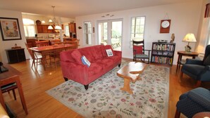 A spacious living room with a red sofa, wooden coffee table, and a floral rug. There is a bookshelf with books and a lamp. A dining area with wooden chairs and a table is visible in the background. - 3 Bedroom house in Cotuit with screened porch and bay view (Barnstable)