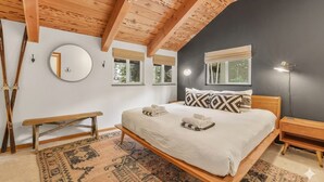 Un dormitorio con techo de madera, una cama con ropa blanca, un espejo redondo y un banco.