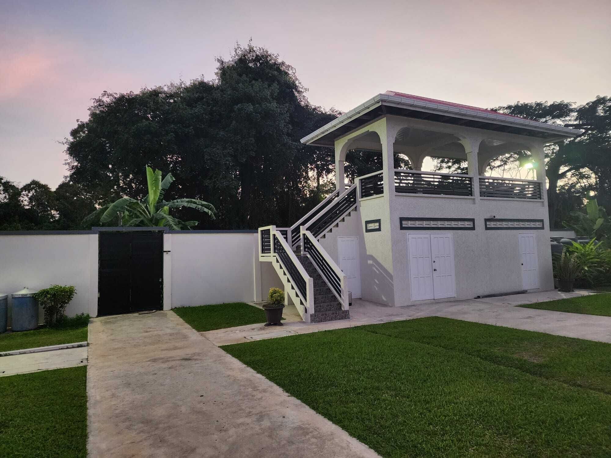 Un espacio al aire libre cubierto con una escalera que conduce a un nivel superior también cubierto, rodeado por un muro blanco bajo y césped verde.